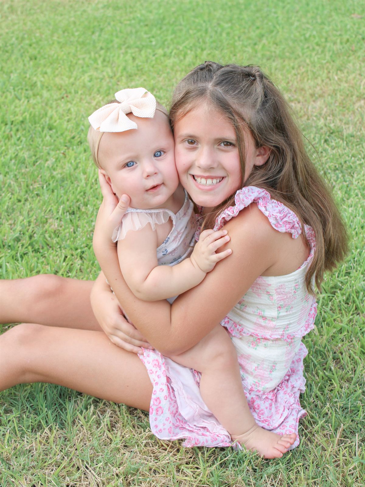 Smiling big sister hugging her baby sister, who wears a cream bow, on the grass